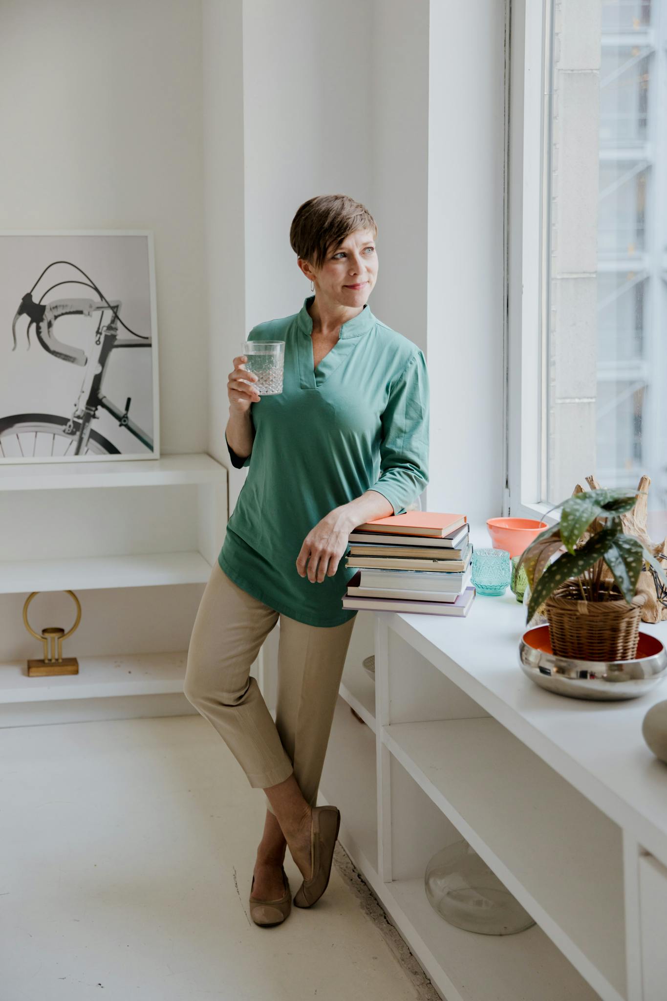 A woman in green stands by a window holding a glass, surrounded by books and indoor plants, exuding calmness.