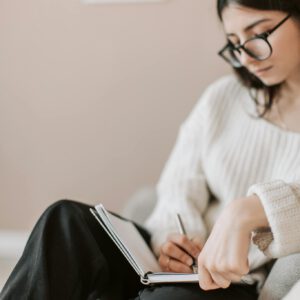 Attentive female wearing eyeglasses and casual outfit sitting barefoot with crossed legs on comfortable couch in modern flat and taking notes in notebook with pen